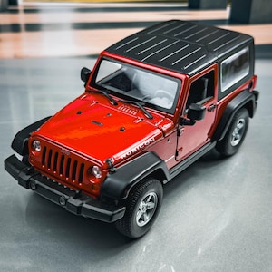 Può includere: Un modello rosso di Jeep Wrangler Rubicon con tetto nero e dettagli neri. L'auto giocattolo presenta una griglia anteriore, fari e ruote. Il modello è esposto su una superficie riflettente.