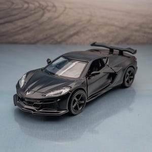 May include: A matte black die-cast model sports car featuring a low-profile aerodynamic body, a rear spoiler, five-spoke wheels, and sharp angular headlights. The vehicle has a sleek, modern design with a prominent front grille and side air intakes.