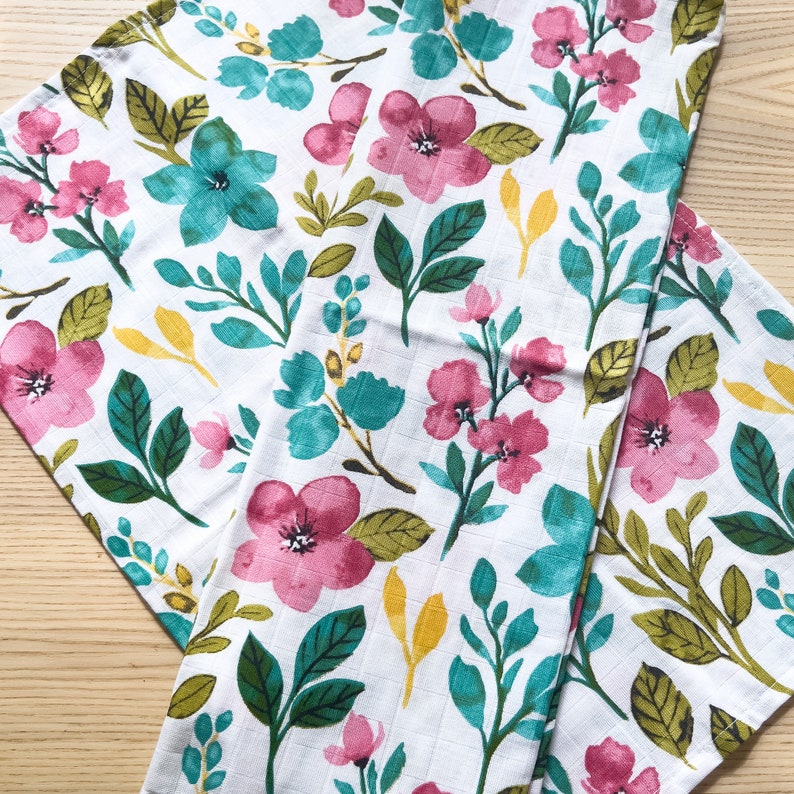 May include: Two white fabric dish towels with a floral pattern. The pattern features pink flowers, green leaves, and teal blue flowers. The towels are folded on top of each other.