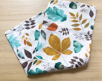 May include: White fabric with a colorful leaf pattern in shades of teal, gold, brown, and orange. The leaves are scattered across the fabric, creating a fall-themed design. The fabric is folded, showing the pattern on both sides.