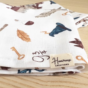 May include: White cotton tea towel with a whimsical pattern of keys, feathers, wands, and a wizard's hat. The towel has a brown label with the text "Heureux Creations".