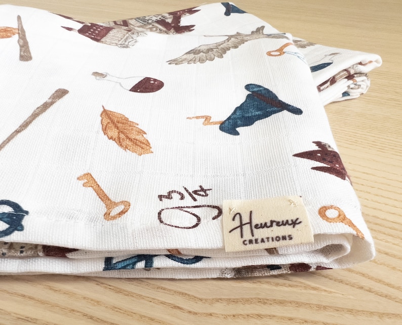 May include: White cotton tea towel with a whimsical pattern of keys, feathers, wands, and a wizard's hat. The towel has a brown label with the text "Heureux Creations".