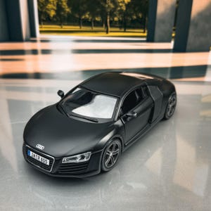 May include: A matte black Audi R8 scale model car featuring a sleek aerodynamic body, detailed headlights, side air intakes, and a front license plate that clearly displays the alphanumeric text IN R 805 on a white background with a blue emblem.