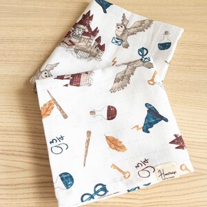 May include: Two white muslin baby burp cloths with a Harry Potter theme. The cloths feature a variety of images including Hogwarts castle, owls, potions, wands, keys, and glasses. The text "3/14" is written on both cloths. The bottom cloth has the text "Hurray Creations" printed on a tag.