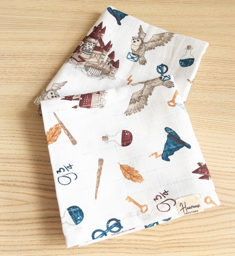 May include: Two white muslin baby burp cloths with a Harry Potter theme. The cloths feature a variety of images including Hogwarts castle, owls, potions, wands, keys, and glasses. The text "3/14" is written on both cloths. The bottom cloth has the text "Hurray Creations" printed on a tag.