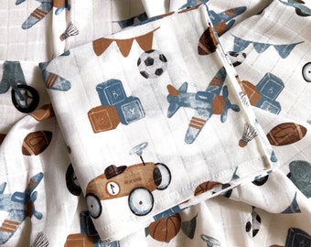 May include: White muslin baby burp cloth with a blue and brown print of airplanes, cars, blocks, and other toys. The fabric is soft and absorbent.