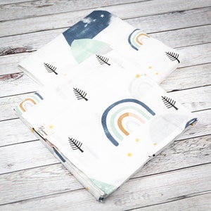May include: Two white muslin swaddles with a rainbow, mountain, and pine tree pattern. The swaddles are folded and stacked on top of each other. The top swaddle has a blue mountain and the bottom swaddle has a rainbow with a yellow, orange, and green color scheme. The text "Handmade Creations" is printed on the bottom swaddle.