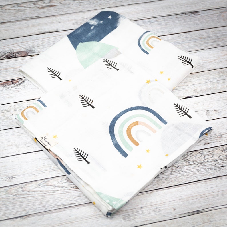 May include: Two white muslin swaddles with a rainbow, mountain, and pine tree pattern. The swaddles are folded and stacked on top of each other. The top swaddle has a blue mountain and the bottom swaddle has a rainbow with a yellow, orange, and green color scheme. The text "Handmade Creations" is printed on the bottom swaddle.