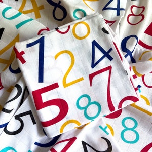 Könnte beinhalten: Eine weiße Musselin-Babydecke mit bunten Zahlen darauf gedruckt. Die Zahlen sind 1, 2, 4, 5, 7 und 8. Die Zahlen sind in Rot, Gelb, Blau und Türkis.