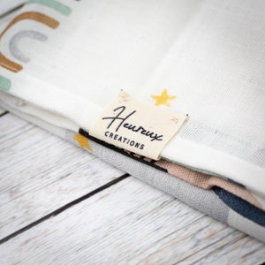 May include: A white fabric with a colorful rainbow design and a small tan label that says "Heureux Creations".