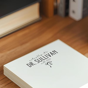 May include: A stack of white square notepads with the text "actually it's DR. SULLIVAN est. 2025" printed in black. The notepads are on a wooden surface, with a keyboard and a plant in the background.