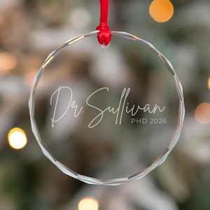 May include: A circular clear glass ornament with faceted edges, suspended by a red ribbon. The ornament features white engraved text that reads Dr. Sullivan PHD 2026 in a combination of script and sans-serif fonts, centred on the glass surface.