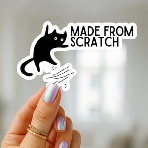 May include: A white sticker with a black cat illustration and the text "MADE FROM SCRATCH". The cat is jumping and appears to be scratching. The sticker is held by a hand with iridescent nail polish and gold rings.