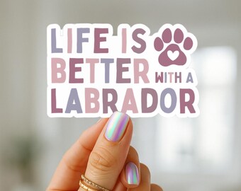 May include: A sticker with the text "LIFE IS BETTER WITH A LABRADOR" in shades of purple and lavender. A paw print with a heart is above the word "LABRADOR". The sticker is held by a hand with iridescent nail polish and gold rings.