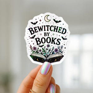 May include: A die-cut sticker featuring the text BEWITCHED BY BOOKS in black lettering. The design includes a crescent moon, small bats, stars, and a central open book with purple and green floral illustrations emerging from the pages.