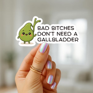 May include: A sticker featuring a cartoon gallbladder with a smiling face and the text "BAD BITCHES DON'T NEED A GALLBLADDER". The sticker is held by a hand with iridescent nail polish and gold rings.