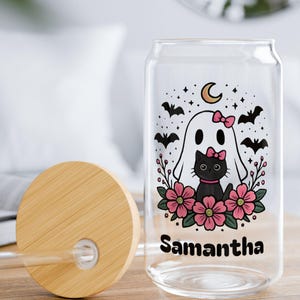 以下が含まれることがあります： 竹製の蓋とストローが付いた、透明なガラス製の缶型タンブラー。タンブラーには、お化け、黒猫、ピンクの花、コウモリ、そして「Samantha」という名前が描かれたハロウィーンをテーマにしたデザインが施されています。