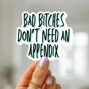 Puede incluir: Una pegatina blanca con el texto "BAD BITCHES DON'T NEED AN APPENDIX" en letras negras con un contorno azul claro. La pegatina está sujeta por una mano con anillos dorados y esmalte de uñas iridiscente.
