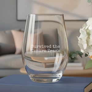 May include: A clear stemless wine glass featuring the white text "Survived the Tenure Track" printed on the front. The glass is positioned on top of a stack of four books with blue, red, green, and yellow covers, resting on a wooden surface.