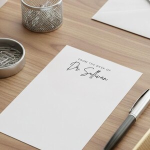 May include: A white notepad with the text "FROM THE DESK OF Dr. Sullivan" in black script. A silver pen, paperclips, and a small succulent in a white pot are also on the wooden desk.