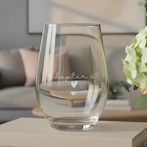 May include: A clear stemless wine glass featuring the white printed text "chapter 60" with a small heart icon below it. The glass is positioned on top of a stack of three books, with a vase of peach and green flowers placed to the right side.
