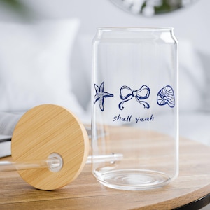 May include: A clear glass can-shaped tumbler with a bamboo lid and straw. The glass is decorated with blue line art of a starfish, bow, and seashell, with the words "shell yeah" below. The tumbler sits on a wooden table.