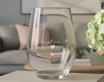 Puede incluir: Una copa de vino sin tallo transparente con el texto impreso en blanco "chapter 35" y un pequeño icono de corazón debajo. La copa está colocada sobre una pila de cuatro libros de tonos claros, con un florero a la derecha.