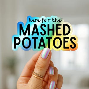 May include: A holographic sticker with the text "here for the MASHED POTATOES" in black letters with a rainbow gradient. The sticker is held by a hand with gold rings and iridescent nail polish.
