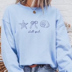Op de afbeelding: Lichtblauw sweatshirt met een marineblauw ontwerp van een zeester, strik en schelp. De tekst "shell yeah" staat eronder.
