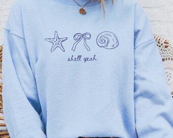 May include: A light blue sweatshirt featuring a starfish, bow, and seashell graphic with the text "shell yeah" in navy blue. The sweatshirt is a crew neck style.