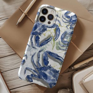 May include: A white and blue iPhone case featuring a watercolor design of blue crabs and green foliage. The case is on a wooden surface with a pen, notebook, and other stationery items.