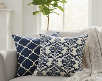 May include: Two decorative throw pillows with navy blue and white patterns rest on a gray sofa. One pillow features a quatrefoil design, while the other has a damask pattern. A beige knitted throw blanket is draped over the sofa.