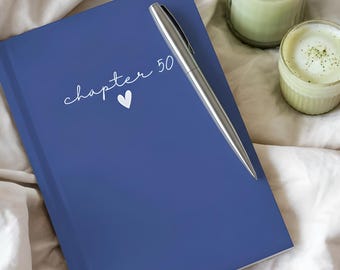 May include: A blue journal with the text "chapter 50" and a heart symbol, with a silver pen resting on top. A green candle in a glass jar and a small cup of matcha latte are also present, all on a white sheet.