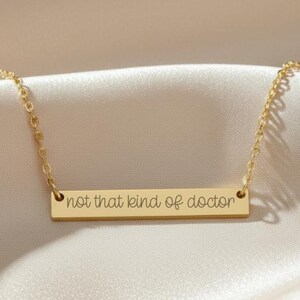 May include: A gold-toned necklace featuring a thin, horizontal rectangular bar pendant. The bar is engraved with the black text 'not that kind of doctor' in a cursive script font. The pendant is suspended from a delicate, matching gold-toned chain.
