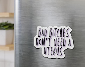 May include: A white sticker with the text "BAD BITCHES DON'T NEED A UTERUS" in black and purple lettering. The sticker is on a silver refrigerator door, with three magnets above it.