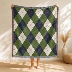 May include: A woven throw blanket with an argyle pattern. The design features green, navy blue, and cream-colored diamonds, accented with red stitching. The blanket has a fringed edge and is held up by a person.