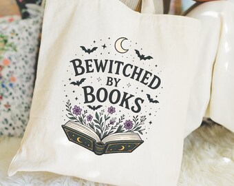 Puede incluir: Una bolsa de tela color crema con un estampado gráfico negro que dice BEWITCHED BY BOOKS. El diseño incluye un libro abierto con flores moradas, pequeños murciélagos, estrellas y una luna creciente, creando una ilustración detallada.