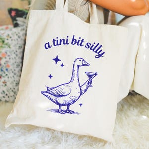 May include: Cream-colored tote bag with a blue illustration of a goose holding a martini glass. The text "a tini bit silly" is arched above the goose. Four blue star shapes are also printed on the bag.