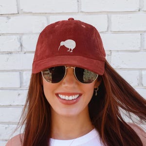 May include: A burgundy corduroy baseball cap featuring a white kiwi bird design. The cap has a curved brim and a six-panel construction. The hat is worn by a person wearing sunglasses.
