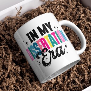 May include: A white ceramic mug featuring the black text IN MY DISSERTATION Era. The word DISSERTATION is printed in alternating vertical stripes of pink, yellow, teal, and blue. Small black star icons are scattered around the bold lettering.