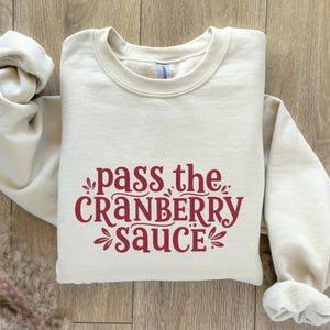 Puede incluir: Sudadera de color crema con la frase "pass the CRANBERRY sauce" en borgoña. El texto está centrado en la parte delantera de la parte superior de cuello redondo y manga larga. La sudadera es de material suave.