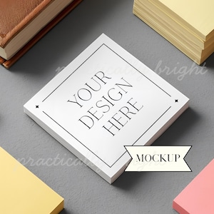 May include: A white square notepad with the text "YOUR DESIGN HERE" in black, framed by a black border. The word "MOCKUP" is in a white banner. Other notepads and a book are in the background.