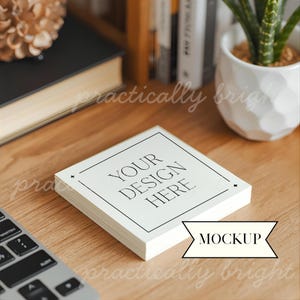 Op de afbeelding: Een stapel vierkante witte kaarten met de tekst "YOUR DESIGN HERE" in een zwart, elegant lettertype. De kaarten liggen op een houten oppervlak, met een toetsenbord en andere kantoorbenodigdheden op de achtergrond. Een "MOCKUP" banner is ook aanwezig.