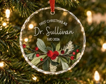 Puede incluir: Un adorno navideño circular de cristal transparente con una cinta roja. El texto dice First Christmas as Dr. Sullivan EdD 2026. Debajo del texto hay una ilustración impresa de hojas de acebo verdes, agujas de pino blancas y bayas rojas.