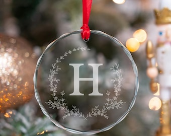 May include: A circular clear glass ornament with beveled edges, featuring an etched letter H in the center surrounded by a delicate leaf wreath design. The ornament is suspended by a thin red ribbon loop for hanging on a holiday tree.