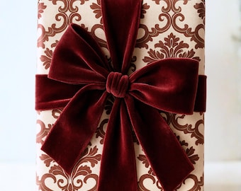 May include: A wrapped gift box with a cream-colored background and a repeating brown damask pattern. A wide, deep red velvet ribbon is tied around the box and forms a large bow on top, creating a luxurious presentation.