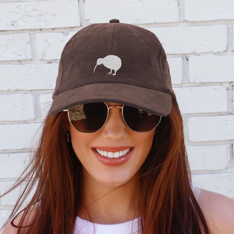 May include: A brown corduroy baseball cap featuring a white kiwi bird design. The cap has a curved brim and a top button. The cap is worn by a person wearing gold-rimmed sunglasses.