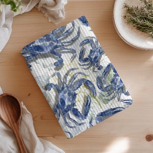 May include: A white and blue patterned kitchen towel featuring watercolor illustrations of blue crabs. The towel has a textured surface and is placed on a light wooden table. A wooden spoon and a white plate are also visible.