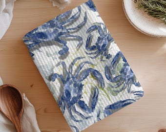Puede incluir: Un paño de cocina con estampado blanco y azul con ilustraciones de acuarela de cangrejos azules. El paño tiene una superficie texturizada y está colocado sobre una mesa de madera clara. También se ven una cuchara de madera y un plato blanco.
