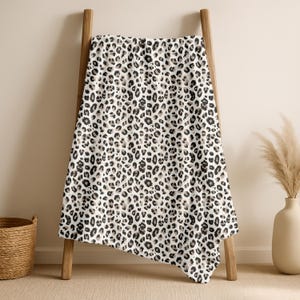May include: A white blanket with a leopard print design, draped over a wooden ladder. The blanket features black and tan spots. A woven basket and a vase with dried plants are in the background.